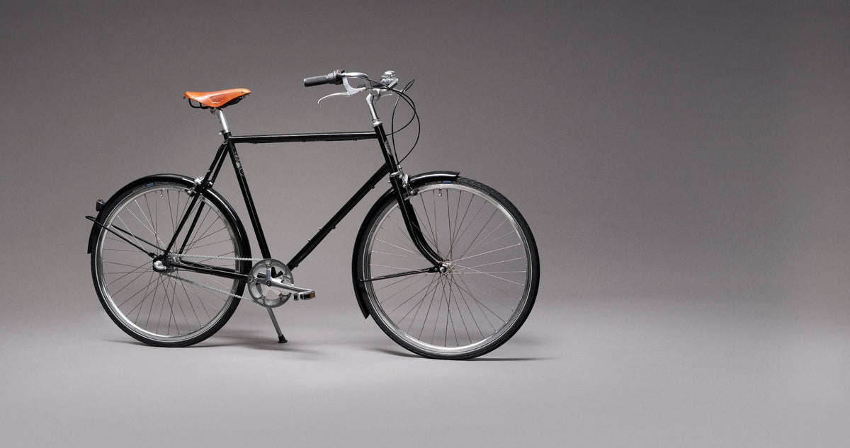 Pelago Bristol, sort bycykel med brun lædersadel, set fra siden på grå baggrund